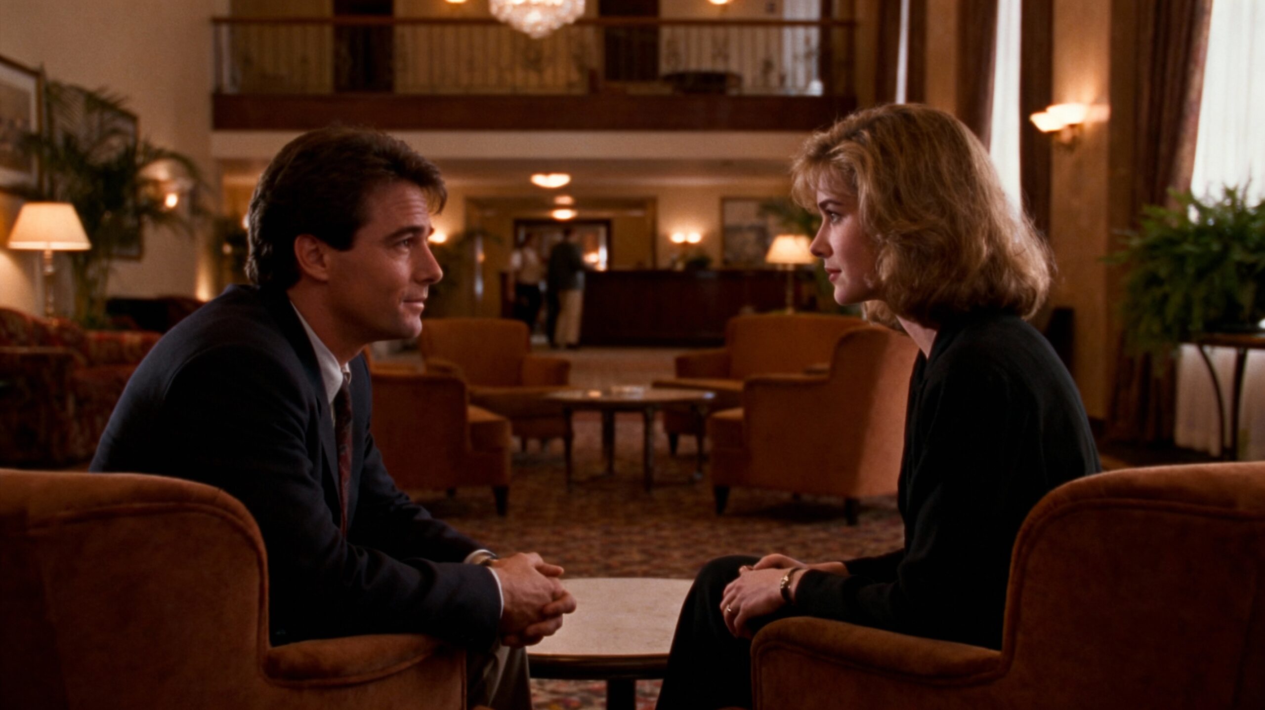 1990s film still of a man and a woman meeting for the first time in a warm hotel foyer. Soft tungsten lighting, slight film grain, muted warm tones, natural imperfections, mid-century furniture, relaxed body language, gentle eye contact. Looks like a frame from a 90s romantic drama, shot on 35mm Kodak film. Cinematic composition, shallow depth of field, atmospheric, understated emotion. --ar 3:2