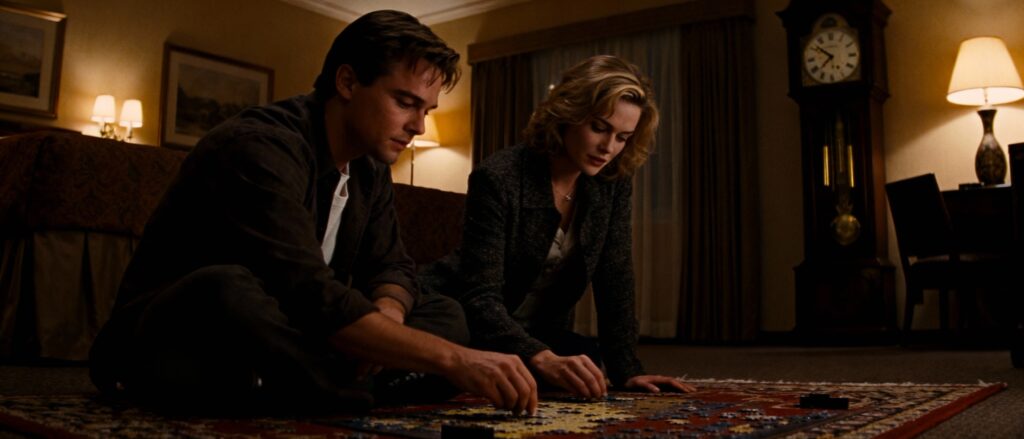 1990s film still of a man and women (quite attractive) working on a jigsaw puzzle, sitting on a rug in a hotel room, grandfather clock in the background - Dark light, warm interior behind them, soft film grain, melancholic tone. --ar 21:9